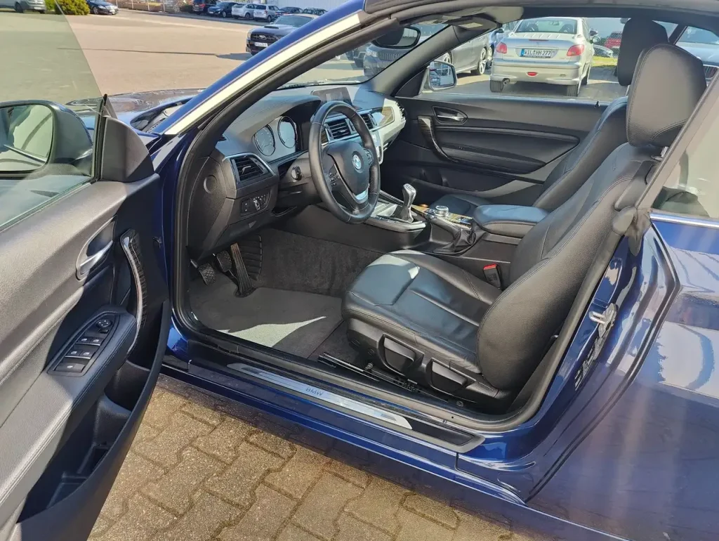 BMW 218 218 i Luxury Line LED Leder Alu Shz PDC, geöffnete Fahrertür, schwarzer Ledersitz, Armaturenbrett mit Lenkrad und Mittelkonsole sichtbar